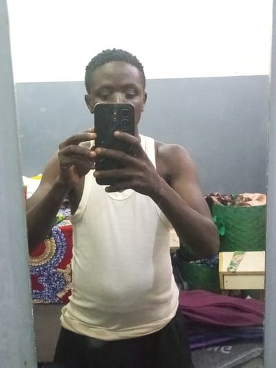 A person is taking a mirror selfie. The person has dark skin, short dark hair, and is wearing a white tank top and black shorts. The location is likely indoors, judging by the plain walls and mirror frame. Behind the person, there's a bed with a colorful patterned blanket, a green woven basket, and some other items covered with a purple cloth on the floor. The person is holding a black smartphone to take the photo. There is nothing in the image to specifically locate it in the city of Rigasa, Nigeria.