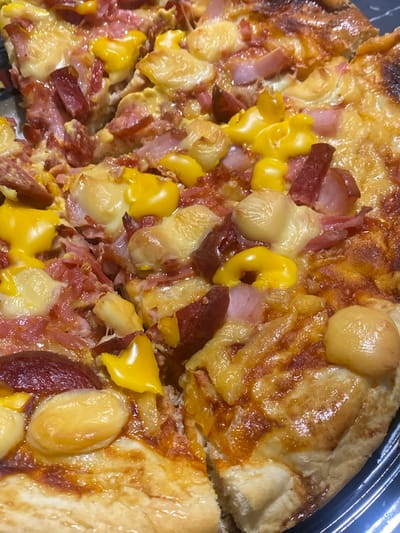 The image displays a close-up, overhead view of a pizza with various toppings. The pizza appears to be freshly baked, with melted cheese oozing over the ingredients. Visible toppings include pieces of what looks like ham or bacon, red onion, and corn kernels. Some of the cheese has a golden-brown, slightly browned appearance, indicating it has been baked to a crisp. The crust is thick and appears to be golden brown. The pizza is cut into several slices, and some of the slices are slightly overlapped, suggesting it is being served from a larger pie. The pizza is placed in a black plastic container, likely a pizza box or takeout tray. The lighting in the image is bright, highlighting the texture and colors of the food. There are no people, specific time of day cues, weather conditions, or visible text in the image. The setting is likely a kitchen or dining area where the pizza is being prepared or consumed.