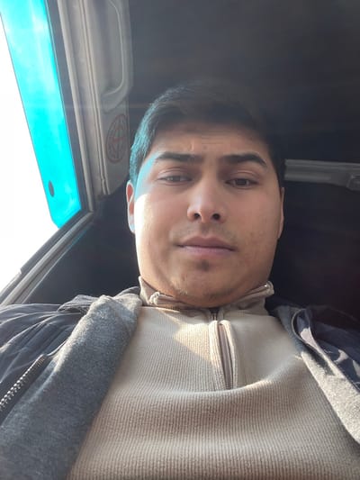 The image captures a male individual seated inside a vehicle, likely in Balykchi, Uzbekistan. The man is positioned centrally, facing the camera from the chest up. He has dark hair and a light complexion, with a small goatee below his lower lip. He is dressed in a beige, ribbed, quarter-zip pullover, over which a dark jacket is worn. A grey fabric element, possibly a collar or hood lining, is visible over the jacket on the viewer's left side.
Strong natural light enters from the left, illuminating the left side of his face and upper body, while the right side is in shadow. To the upper left of the subject, a vehicle window shows a bright, featureless sky, appearing with a blue-green tint. A light-colored grab handle is affixed to the vehicle's interior trim above the window. Adjacent to the grab handle, a circular "NO SMOKING" decal is visible, featuring a red border, a diagonal line, and a black cigarette icon. The vehicle's ceiling is dark. No specific actions or interactions are depicted beyond the subject looking at the camera.