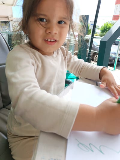 In Ümraniye, Turkey, a young girl is pictured sitting at a table outdoors, possibly at a cafe, as suggested by the presence of a grey upholstered chair and a table surface. She is wearing a light beige long-sleeved shirt and light beige pants. The girl is looking towards the camera with a slight smile and her mouth open. Her hair is dark and appears windswept. In front of her, she is holding a green marker and drawing on a piece of white paper. The background shows a street scene with cars, trees, and buildings, indicating an urban setting. A metal pole with directional signs can be seen to the right of the girl.