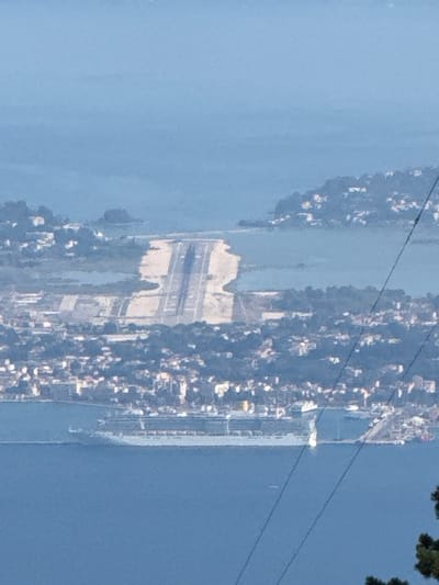 The image shows a panoramic view of the city of Kassopaia Municipal Unit, Greece, from an elevated vantage point. In the foreground, a large body of water, likely the Ionian Sea, dominates the lower portion of the frame. A cruise ship is visible on the water, its white hull and numerous decks clearly discernible. Further inland, an airport runway is prominent, appearing as a long, paved strip cutting through the landscape. The surrounding area is characterized by a dense urban sprawl, with buildings clustered together on hillsides overlooking the sea and the airport. Lush green vegetation is interspersed with the urban development. In the far distance, a hazy blue sky meets the horizon. The image is captured in daylight, with clear visibility, though a slight haze softens the distant features.