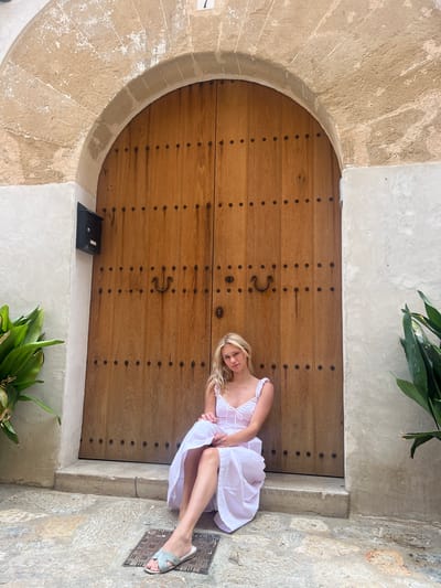A young, blonde woman in a white sundress sits on the stone steps of an arched wooden doorway in the town of Pollença, Spain. The doorway, made of vertical wooden planks and studded with dark metal accents, features a large, U-shaped metal handle and a keyhole. Above the doorway, a rough-hewn stone archway is visible, with a numeral '7' appearing at the top. To the left of the door, a black mailbox is mounted on a white plastered wall, and a lush green plant with broad leaves grows nearby. To the right, another green plant with similar leaves is partially visible. The ground in front of the doorway is paved with textured stone, and a metal grate is set into the paving stones near the woman's feet. The woman is looking towards the camera with a slight smile, her legs crossed, and her hands resting on her lap. The overall impression is one of a serene and picturesque scene in a historic Mediterranean town.