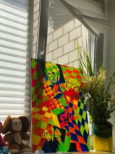 The image depicts a colorful abstract painting with stylized faces rendered in a mosaic-like pattern of diamond shapes. The painting is set on a windowsill next to a plush toy and a vase of greenery. Sunlight streams in from the right, illuminating the scene. The windowsill appears to be in an urban setting, possibly in the city of Cherven, Belarus, given the context. The overall impression is one of domestic tranquility, with artistic expression and natural elements coexisting in a sunlit space.