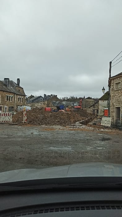 An outdoor scene in Florenville, Belgium, depicts an active construction or demolition site under an overcast gray sky. The foreground shows a wet, uneven paved surface with visible puddles, framed at the bottom by a vehicle's dark dashboard. Centrally, a large mound of reddish-brown earth, rubble, and assorted debris dominates the mid-ground. An orange and red hydraulic excavator is visible behind the debris pile, positioned towards the right, along with a darker blue construction vehicle further back on the left. Red and white striped construction barriers are erected on both the left and right sides of the debris field, marking the perimeter of the work area. On the far right, a narrow excavated trench runs parallel to a stone building with a decorative black street lamp attached; an orange portable utility unit is visible nearby. To the left and in the background, multiple traditional stone buildings with dark slate roofs and chimneys are present. Utility poles and overhead wires are visible on the right side of the frame. No individuals are depicted.