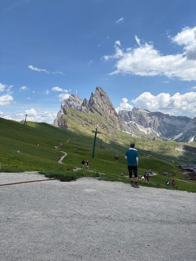 The image depicts a mountainous landscape in Santa Cristina Gherdëina - St. Christina in Gröden - Santa Cristina Valgardena, Italy. In the foreground, a person stands facing away from the camera, looking at the expansive mountain range. The person is wearing a teal t-shirt, black shorts, and hiking boots, with a white towel draped around their neck. The ground in the immediate foreground is covered in gravel.
A winding, paved path leads down the green, grassy mountainside, dotted with yellow wildflowers. Several other people are visible on the path and scattered across the grassy slopes, some walking, others resting. Ski lifts are present on the left side of the slope, extending upwards. The background is dominated by imposing, jagged grey mountains with patches of snow on their peaks, set against a bright blue sky with scattered white clouds. The overall scene suggests a hiking or recreational area in the Dolomites during a clear day.