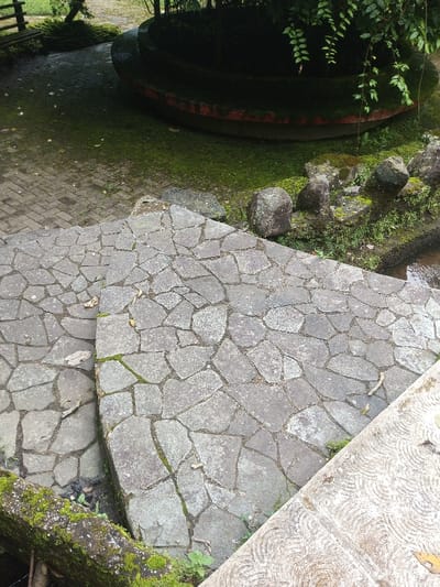 The scene depicts an outdoor landscaped area in Kanand, India. In the immediate foreground and midground, a pathway and steps are constructed from irregularly shaped, light grey flagstones, exhibiting visible moss growth within cracks and along edges. To the immediate right, a section of a light-colored concrete structure with a concentric circular pattern is visible. Below the flagstone path, a narrow water channel with dark water is present, bordered by multiple dark, moss-covered rocks and small green plants. In the background, a large, dark circular stone structure with a reddish-brown base is situated on a patch of bright green mossy ground. This structure is heavily shaded by overhanging green foliage. To the left of the circular structure, a brick-paved area extends into the distance, transitioning to additional green ground cover and distant vegetation. No individuals or active subjects are present.