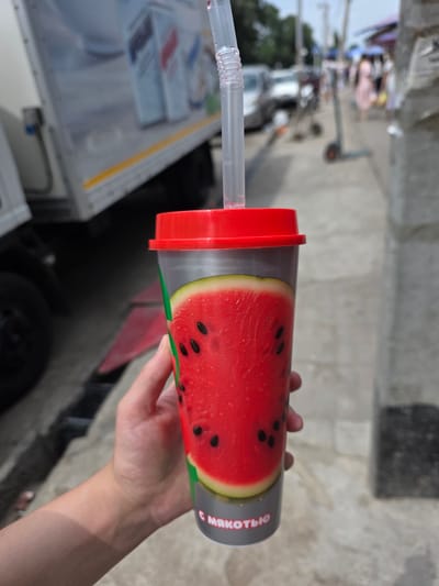 A close-up vertical shot shows a person's hand holding a plastic cup with a watermelon design. The cup has a red lid and a clear straw. In the background, a truck is parked on the left side of the image. The right side of the background shows a street scene with people walking and shopping under umbrellas. The setting appears to be a street market in the city of Talgar, Kazakhstan. The overall lighting suggests it is daytime.