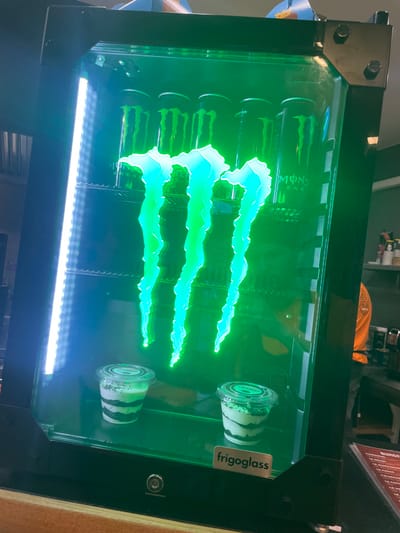 The scene depicts the interior of a commercial establishment, likely a cafe or snack bar, located in Holtzheim, France. In the foreground, a black-framed "frigoglass" display refrigerator stands on a light brown wooden counter. The refrigerator is illuminated by a prominent neon green Monster Energy logo, which appears to be part of the unit's design. A vertical white LED light strip runs along the inner left side of the refrigerator door.

Inside the refrigerator, on the upper shelf, are multiple black cans labeled "MONSTER ENERGY." On the bottom shelf, two clear plastic cups contain layered desserts, featuring alternating white and dark strata, topped with green-colored crumbles. The refrigerator features metallic bolted corner reinforcements and a central lock on its lower front panel.

In the blurred background, a person wearing an orange long-sleeved shirt is partially visible to the right. Behind them, shelves with various items, including cups and bottles, can be seen. A dark menu stand with visible text is present on the right edge of the frame. The overall environment behind the refrigerator is dimly lit.