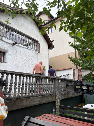 This is a candid, outdoor photograph taken in Konjic, Bosnia and Herzegovina. The image is oriented vertically, with the camera looking upwards towards a building and overhanging tree branches.
In the foreground, a stone balustrade with decorative pillars runs along a balcony. On this balcony, a bald man, shirtless and wearing dark trousers, is visible from the back. He appears to be leaning over the railing. Further along the balcony, a person in a dark blue t-shirt is partially visible. A stack of black vests, possibly safety equipment, is seen on a higher level of the building. In the background, there is a white building with a brown-tiled roof and a tan-colored section of the building. The scene suggests a residential or multi-story building with outdoor living spaces.
The activity appears to be casual, with the shirtless man perhaps looking over the edge or interacting with someone below. The presence of the vests might suggest some form of activity related to water sports or rescue, given the context of Konjic being known for its river activities.
Notable details include the texture of the white stucco wall and the intricate design of the balustrade. The lighting suggests it might be daytime, possibly overcast given the soft shadows. There is a water bottle with a blue cap on the balcony. No visible text is present in the image.