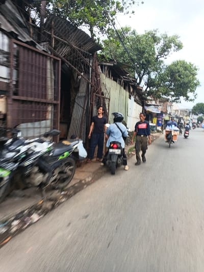 The image presents a street scene in Dayeuhkolot, Indonesia, characterized by a mix of worn commercial or residential structures and ongoing street activity. On the left, a row of buildings constructed primarily from corrugated metal sheets, exhibiting rust and weathered appearance, runs parallel to the asphalt road. One building features a dark, open entrance with a rusted metal gate partially visible. Two motorcycles are parked on the muddy shoulder adjacent to these structures; the closest is black with green accents.
Three individuals are prominent in the foreground. Standing near the open entrance on the left is a male with dark hair and a beard, wearing a black t-shirt and dark pants, looking upwards and to the right. To his right, a person wearing a light blue long-sleeved shirt, dark pants, and a black motorcycle helmet is seated on a stationary black scooter motorcycle, facing away from the camera. Further to the right and slightly ahead, another male, wearing a dark blue t-shirt with a white logo and dark cargo pants, walks forward along the roadside.
The street extends into the background where more buildings are visible, suggesting a denser urban area. A person on a bicycle with a loaded cart is also visible further down the road, along with other distant figures and vehicles. Numerous black electrical or communication wires are strung overhead, and green tree foliage is visible above the structures and in the distance against an overcast sky. The road surface appears grey and worn, with some debris present on the immediate roadside.