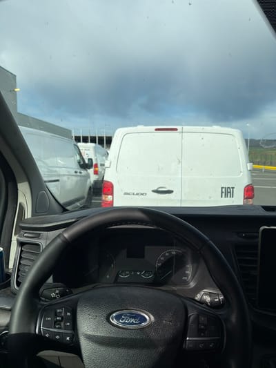 The image, taken from inside a Ford vehicle, shows a dashboard and steering wheel, as well as a view through the windshield. Outside, there's a Fiat Scudo van directly in front of the Ford. Other white vans are visible in the distance behind the Fiat. The environment appears to be a parking area or industrial zone under an overcast sky. There's no indication the location is Goßfelden, Germany.