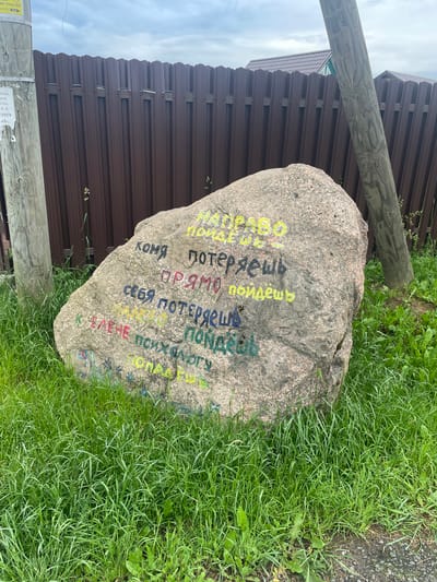 The image depicts a large, grey boulder covered in multi-colored graffiti, situated in a grassy area next to a brown, metal fence and a wooden utility pole. The graffiti on the boulder appears to be written in Russian. In the background, two houses with tiled roofs are visible. The sky is overcast. The scene is outdoors, with the boulder being the central subject, adorned with handwritten text and symbols. The surrounding environment consists of grass, a fence, and a utility pole, suggesting a semi-rural or residential setting. There are no people visible in the image.