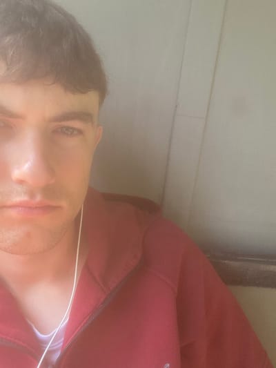 A close-up, rotated view shows a person's face partially in frame. They have short dark hair, light skin, and are wearing headphones. They are wearing a red zippered jacket. The background consists of a light-colored surface with horizontal lines, possibly a wall. The photo's origin in Matyashivka, Ukraine is not determinable from the image content.