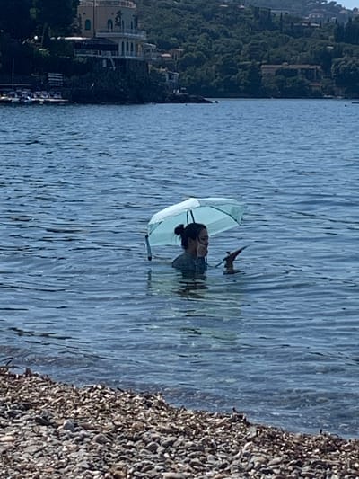 In the city of Pozzarello, Italy, a person stands in the water holding a light blue umbrella. The person is submerged up to their chest and appears to be holding a phone or similar device in their right hand. The background features a shoreline composed of pebbles, with a body of water extending towards a distant coastline characterized by dense green foliage and buildings. The sky is clear and blue, suggesting a sunny day.