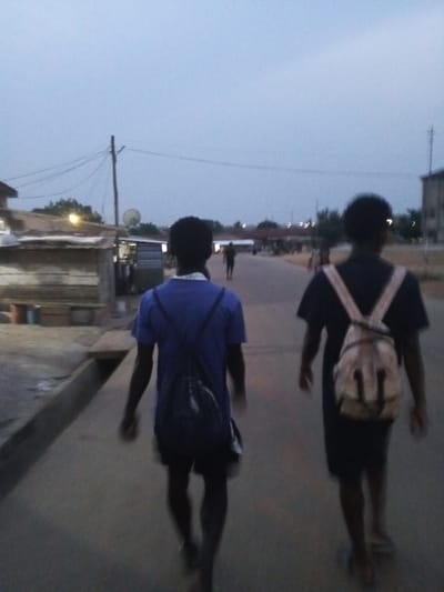 The image depicts an evening scene in Kwamo, Ghana, with two individuals walking away from the camera down a street. The person on the left wears a blue shirt and a dark drawstring backpack, while the person on the right is in a dark shirt and carries a lighter, possibly beige or tan, backpack that appears to have a subtle design. Their figures are seen from behind, partially silhouetted against the dim light.

The time of day is clearly dusk or early evening; the sky above is a pale grey-blue, but the street level is dark, and artificial lights are visible in the distance and near the buildings on the left. The road appears paved, and a concrete gutter runs along its left edge. On the left side of the street, there are several simple, low-rise structures, possibly market stalls or small shops, with some lights glowing. A utility pole with numerous overhead wires and a satellite dish is also visible on this side. In the background, other people can be seen walking further down the road, and the outlines of distant buildings are faintly discernible against the twilight sky. No discernible text is visible.