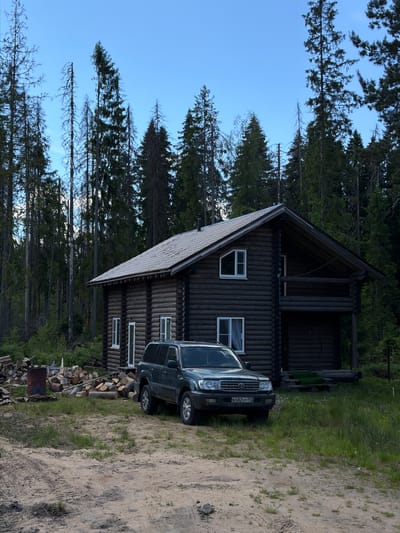 This image displays a two-story log cabin situated within a dense coniferous forest clearing under a clear blue sky, in Lebyazh'e, Russia. A dark green Toyota Land Cruiser SUV (100 series) with Russian license plate B582XP 147 is parked on a sandy-dirt clearing directly in front of the house.
The log cabin is constructed of dark-stained horizontal logs and features a gabled roof covered with light gray corrugated metal. Visible windows include two white-framed rectangular windows on the front facade, one on each story, and another white-framed, irregularly shaped window on the upper story of the left side. A covered wooden balcony with log railings extends from the upper story on the right side of the house, above a dark, recessed lower area. A dark-colored chimney protrudes from the left side of the roof.
To the left of the house, a significant pile of stacked cut firewood and timber is present on the ground, next to a rusty red metal barrel. The immediate foreground consists of a sandy-dirt surface with distinct vehicle tire tracks and scattered patches of green grass. The surrounding environment is characterized by a thick forest composed of numerous tall, slender evergreen trees, some appearing vibrant green while others exhibit bare or dead branches. The sky above is predominantly light blue with minimal cloud cover.