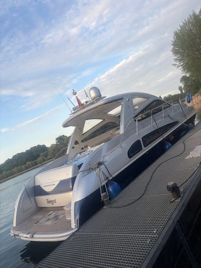 The image shows a yacht named "Target" docked at a marina in Germersheim, Germany. The yacht is predominantly white with navy blue accents. It is moored alongside a metal-grate pier. On the pier, there's a mooring post. The sky is visible in the background, partially obscured by the yacht's structure and trees lining the shore in the distance. The water is calm. Flags are on the yacht's mast.