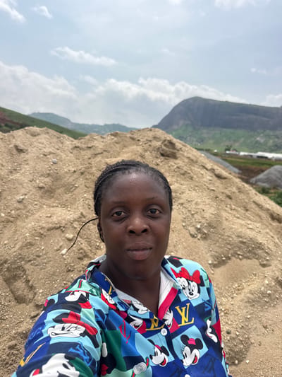 A medium shot captures a Black woman from the chest up, taking a selfie outdoors. She is positioned in front of a large, sloping pile of sand. In the background, distant mountains rise under a cloudy sky, with some green vegetation visible on their slopes and a few structures in the mid-ground. The woman has dark skin, dark hair braided into two strands, and is wearing a colorful shirt adorned with images of Minnie Mouse and the Louis Vuitton logo. Her expression is neutral. The scene appears to be a construction or material site, possibly in the vicinity of Jawu, Nigeria, given the mountainous terrain and open landscape.
