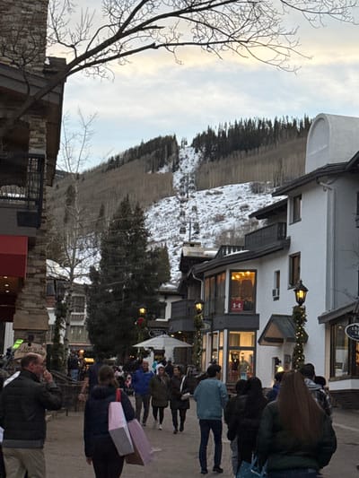 A street scene displays people strolling and shopping, with buildings lining the walkway. Christmas lights adorn the storefronts, adding a festive touch. In the background, a snow-covered mountain rises, complete with a ski lift. A bare tree stands on the left, silhouetted against the sky. The overall ambiance suggests a bustling village nestled in a picturesque mountain setting.