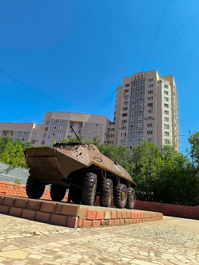 A brown armored personnel carrier is displayed on a brick pedestal outdoors, with a modern, multi-story building and trees in the background under a clear blue sky. The APC, positioned at a low angle perspective, faces away from the viewer, revealing its six large, treaded tires and the rear portion of its hull, which shows some signs of weathering and damage. The pedestal is constructed from red bricks and is situated on a paved area with a mosaic-like pattern. The building behind the APC is a tall, contemporary structure with numerous windows and balconies, suggesting an urban environment. Lush green trees partially obscure the lower sections of the building, indicating a green space within the city. Power lines are visible stretching across the sky.