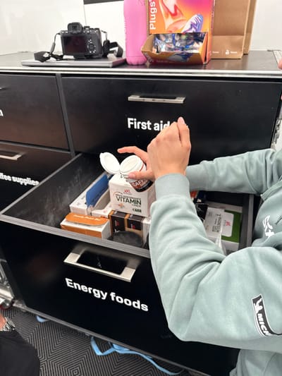 The image shows a person's arm and hand reaching into an open drawer of a black metal cabinet. The drawer is labeled "First aid" on the top and "Energy foods" on the bottom. The person is holding a small, white container that is partially open, revealing its contents. Inside the drawer, there are various products, including boxes labeled "New Performance Vitamin" and "Magnesium". The cabinet is located in Stavelot, Belgium, at coordinates 50.441 latitude and 5.969 longitude. The cabinet is located near an assortment of objects, including a camera, a pink water bottle, and an assortment of candies. The scene is located in a room that appears to be an indoor setting. The person is wearing a light blue sweater with a patch on the sleeve.