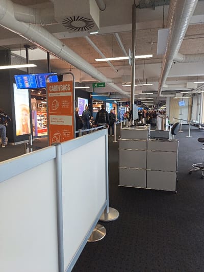 The image depicts an indoor airport concourse, specifically within EuroAirport Basel Mulhouse Freiburg, located in Saint-Louis, France. The scene is brightly lit by overhead linear fluorescent fixtures and spotlights on an industrial-style ceiling featuring exposed white circular air ducts and ventilation grilles. The floor is covered with a dark grey patterned carpet.

In the immediate foreground, a temporary, freestanding white partition wall with silver framing and circular bases extends across the left side of the frame. To its right, an orange, rectangular sign dominates the mid-ground, displaying "CABIN BAGS" in large white text. Below this, it illustrates and describes baggage policies: "All customers: 1x SMALL UNDERSEAT CABIN BAG" with a backpack icon, and "Customers with Speedy Boarding: 1x SMALL UNDERSEAT CABIN BAG" with a smaller bag icon.

Behind the cabin bag sign and to the left, a retail space is visible, featuring a large illuminated display screen showcasing a woman's face, likely an advertisement. Above this, a blue digital flight information display board is active. To the right of the cabin bag sign, a modular grey service counter or storage unit, composed of six cube-like sections with thin metallic frames, is positioned. A black office chair on wheels with a silver base is partially visible on the far right, beside the counter.

Multiple retractable belt barriers and stanchions delineate pathways within the concourse. In the background, numerous individuals, approximately 15-20, are observed walking and standing. Most are dressed in casual attire, carrying backpacks or bags, consistent with air travelers. The concourse extends into the distance, revealing more digital display screens, a bright blue-lit structure, and a green exit sign with a running figure symbol mounted overhead. People appear to be moving through the area, heading towards unseen gates or checkpoints.