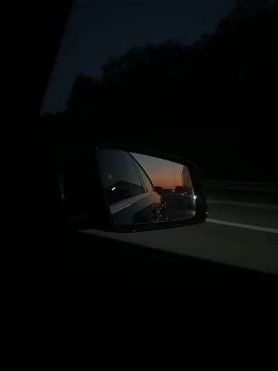 The image depicts a view from inside a car at dusk. The side-view mirror is prominently displayed, reflecting the sky with a gradient of orange and dark blue hues, indicative of sunset. The road is visible in the reflection, with a white line marking the lane. Beyond the mirror, the dark silhouettes of trees are discernible against the twilight sky. The overall scene suggests a journey on a highway, possibly in or around Elsdorf, Germany, given the context. There are no visible people or specific landmarks that would pinpoint the exact location within Elsdorf. The environment is characterized by low light conditions and the speed of travel, implied by the slight blur in the reflected scenery.