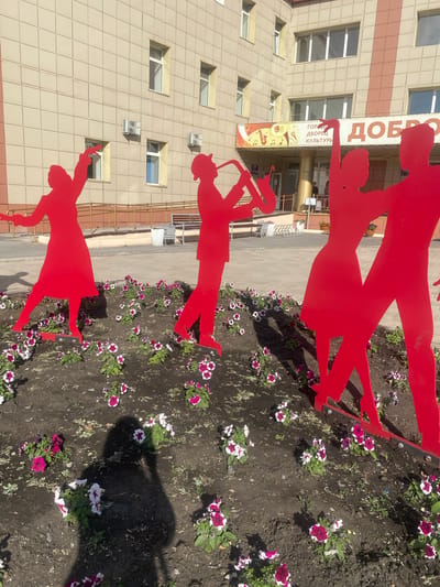 This media file captures a bright, sunny day in Achinsk, Russia, focusing on a flowerbed adorned with red metal silhouettes of dancing figures in front of a modern building. The building, with its cream-colored tiled facade and numerous rectangular windows, suggests a public or commercial space.
The most prominent subjects are the vibrant red silhouettes, depicting at least three pairs of people in dynamic poses, appearing to dance or engage in a lively activity. These silhouettes are arranged vertically, with one pair at the top, another in the middle, and a third at the bottom. Their red color creates a striking contrast against the muted tones of the building and the dark soil of the flowerbed.
The flowerbed itself is filled with blooming petunias in shades of pink and white, adding a touch of natural beauty to the urban landscape. The sunny weather is evident from the strong shadows cast by the silhouettes and the bright illumination of the scene.
A banner with Russian text is visible on the building, partially obscured by the silhouettes. The text includes "ДОБРО" (Dobro, meaning "good" or "kindness") at the top, and below it, "ГОРОДСКОЙ ДВОРЕЦ КУЛЬТУРЫ" (Gorodskoy Dvorets Kultury, meaning "City Palace of Culture"). This inscription clearly indicates the location as a cultural center.
The overall impression is one of festivity and community, with the dancing figures symbolizing joy and shared experiences, set against the backdrop of a cultural institution. The clear sky and abundant sunshine suggest a pleasant time of day, likely midday, suitable for outdoor activities and public gatherings.