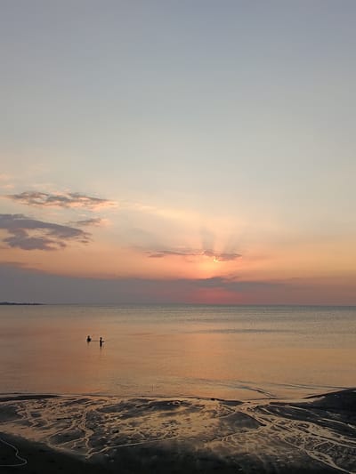 This media file captures a serene sunset scene over the ocean at Jangkar, Indonesia. The sky is a gradient of soft blues and oranges, with clouds tinged pink and gold by the setting sun. Rays of light pierce through a cluster of clouds, illuminating the sky with a warm glow.
The ocean is calm, reflecting the colors of the sky, with a gentle ripple on its surface. In the shallow waters near the shore, two silhouetted figures are visible, appearing to be wading or fishing with long poles. Their presence adds a sense of human activity to the vast seascape.
The foreground consists of a sandy, wet shoreline, marked by intricate patterns of dried sand and water channels, suggesting the ebb and flow of the tide. In the distance, a long pier extends into the water, with a few structures visible at its end. The overall atmosphere is peaceful and tranquil, evoking a sense of quiet contemplation as the day draws to a close. The time of day is clearly sunset, and the weather appears to be clear and calm. There is no visible text in the image.