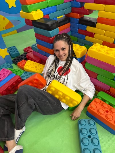 An overhead shot captures a young woman lying playfully on a vibrant green mat, surrounded by and leaning against an array of oversized, colorful building blocks. The blocks, resembling large LEGO bricks, are made of various materials and come in numerous bright hues including yellow, red, blue, green, orange, pink, black, and light blue. Some blocks feature typical stud designs, while others are smooth or have softer, rounded edges.
The woman has her head slightly raised and is smiling broadly at the camera. She has long, dark and light braided hair and is wearing a white short-sleeved t-shirt with a partially visible red heart graphic, dark grey baggy pants, and white socks with blue and white sneakers. The setting appears to be an indoor play area or creative space, possibly designed for children, given the large-scale blocks and star-shaped cutouts visible on the lower left.
Notable text includes "ESDA" and "Made in Germany" embossed on a light blue block. On her white sock, the phrases "WE ARE WILD AND FREE" and "LIVE TOGETHER WITH NATURE" are visible. The overall mood is cheerful and relaxed. The photograph is taken in Korobovo, Russia.