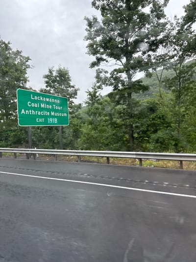 A green highway sign with white lettering reads "Lackawanna Coal Mine Tour Anthracite Museum" and "EXIT 191B". The sign is positioned in front of a dense forest of green trees under a cloudy, overcast sky. A wet asphalt road with a white lane marker stretches across the foreground, indicating recent rain. A metal guardrail runs parallel to the road, separating it from the wooded area behind. The overall scene is that of a roadside view in a rural or semi-rural area, likely in South Abington Township, Pennsylvania, given the mention of Lackawanna County on the sign.