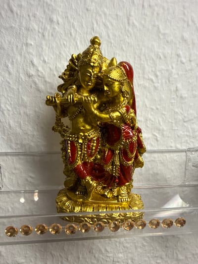A gold-colored statue of Radha and Krishna stands on a clear plastic shelf against a white, textured wall. The statue depicts Krishna playing a flute, with Radha standing beside him, her arm around his shoulder. Both figures are adorned in red and gold clothing with intricate detailing. The shelf itself is decorated with a row of small, clear rhinestones. The lighting appears to be artificial, casting some reflections on the shelf and the statue. This image was taken in Velbert, Germany.