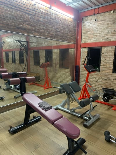 The image shows the interior of a gym located in Kecamatan Kartasura, Indonesia. The gym is equipped with various exercise equipment, including two hyperextension benches, one of which is in the foreground and appears to be manufactured by Olympic, and one of which is reflected in the large mirror on the wall. The room is decorated with red brick walls and a wooden floor. There are two windows and one large mirror covering most of the back wall. The lighting is provided by fluorescent tubes mounted to the ceiling. The gym is empty, with no people present.