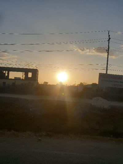 The image shows a sunrise or sunset over a landscape. Silhouetted power lines and a building frame appear on the left. A sign with faded text and numbers is visible at the top. The bright sun casts a glare, illuminating a field or undeveloped area. The right side features a dark road or path with greenery. The overall scene is hazy and the image is rotated, with the horizon running vertically.