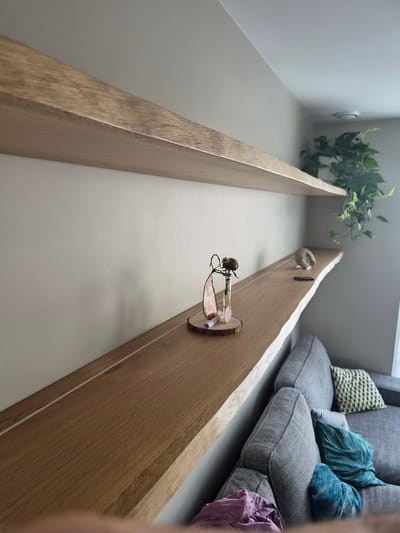 The image depicts an interior scene, likely a modern living space, with a focus on a long, wooden trough-like structure. This structure appears to be built into a wall or furniture, and runs horizontally across the frame. On the trough, a small, decorative item is visible, consisting of a glass vial containing a dried flower or bud, attached to a small wooden disc with twine. Another decorative element, possibly a small burlap ball, is further down the trough.
In the background, to the right, a grey sofa with several decorative cushions in shades of teal and yellow is visible. A piece of purple fabric, perhaps clothing, is draped over the edge of the sofa. To the left of the trough, a green plant cascades over the edge of its container. The overall impression is one of a carefully curated and minimalist interior. The lighting suggests it could be daytime, with natural light coming from an unseen source, possibly a window. No people are visible in the image. The location context of Schengen, Luxembourg is provided but not directly evident from visual cues within the image itself.