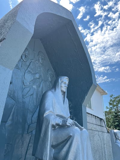 This media file shows a close-up, low-angle shot of a memorial sculpture in Takhtamukai, Russia. The sculpture appears to be made of a dark, textured material, possibly concrete or painted metal, and depicts several figures. In the foreground, a large, silver-toned statue of a person in a hooded garment is visible, their head tilted upwards and their gaze directed towards the sky. The person is holding an object in their hands.

Below and behind the foreground figure, a bas-relief depicts multiple figures of soldiers, each wearing helmets and appearing to be lying down or in repose. Their faces are sculpted with varying degrees of detail, suggesting a somber or respectful tone. The sculpture is set against a backdrop of a bright blue sky with scattered white clouds, indicating a clear day. The sun is casting shadows, suggesting it is daytime. In the upper right portion of the image, the edge of a white building and some green trees are visible, providing a glimpse of the surrounding environment. The overall mood conveyed by the sculpture and the bright sky is one of remembrance and perhaps hope. No visible text is present in the image.