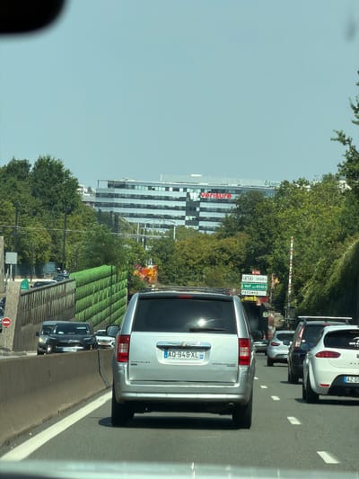 The image shows a view from a vehicle traveling on a multi-lane road, likely a highway or major thoroughfare, in Antony, France. The road is lined with trees and features a large, modern building in the background. The building has a repeating pattern of windows and appears to be an office complex, with "Groupon" and "verisure" signage visible on its facade.

Several cars are visible on the road, indicating traffic. In the foreground, the rear of a silver minivan is prominent, displaying the license plate "AQ-949-XL". Another car, a dark sedan with license plate "GS-244-YN", is ahead of the minivan. Further ahead, other vehicles, including a white car with license plate "AZ-D", are also in view.

Green, textured barriers or structures are present alongside the road, possibly noise barriers or architectural features. Road signs are visible, one indicating "N166" and distances to "L'HAY LES ROSES" and "FRESNES". The sky is a clear, light blue, suggesting daylight, likely daytime under fair weather conditions. The perspective and the presence of vehicles in motion suggest the photograph was taken from a moving car. The overall scene conveys a typical urban or suburban highway environment.