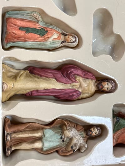 The image shows a nativity scene figurine set, likely made of porcelain or ceramic, displayed within a beige, molded plastic packaging. The figures are arranged in separate compartments.
The top compartment contains a reclining female figure with a pinkish-orange robe draped over a pale blue garment. A dark green object, possibly representing swaddling clothes or a bundle, lies across the robe. The figure's head is partially visible on the right, with painted facial features including closed eyes and dark hair.
The middle compartment holds a reclining male figure. He wears a pale yellow garment and a deep red cloak draped over his shoulders and body. His head is turned to the right, revealing painted facial features with open eyes and a beard.
The bottom compartment shows another reclining male figure wearing a brown garment over a pale blue undershirt. He is holding a small white lamb with a textured wool surface. His head is turned to the right, showing painted facial features with an orange beard and open eyes.
A partial view of a fourth figurine is visible in a compartment on the far right, showing a green garment and part of a red object.
The overall scene is indoors, within the packaging. There is no indication of the city of Carlow, Ireland, or any urban environment.