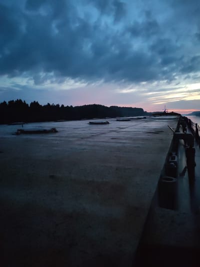 The image shows a flat, concrete surface, likely a dock or pier, stretching into the distance. The surface appears wet and textured, with some debris or irregularities. On the left side of the image, a dark line of trees forms the silhouette of a distant forest against a dusky sky. The sky itself is dramatic, with dark, heavy clouds occupying the upper portion, transitioning to lighter hues of pink and orange near the horizon. The right side of the pier features industrial elements, including what appear to be large metal rings or tires stacked vertically, and metallic structures. A railing runs along the edge of the pier, with a body of water visible beyond it. In the far distance, a faint light or structure is visible on the horizon. The overall scene is one of twilight or early dawn, with subdued lighting and a moody atmosphere. The location in Badozhskoi Pogost, Russia, suggests a potentially remote or industrial waterfront setting. There are no visible people in the image.