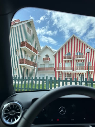 The image displays a daytime scene in Costa Nova, Ílhavo, Portugal, viewed through the windshield of a vehicle. The top portion of a dark dashboard, an air vent with metallic trim on the left, and a blurred steering wheel with a Mercedes-Benz logo are visible in the foreground. The sky above is blue with scattered white, cumulus clouds.
In the mid-ground, a green picket fence spans horizontally, bordering a well-maintained patch of green grass. Beyond the fence, three distinct, multi-story buildings with pitched roofs are visible.
The building on the left features a facade with alternating vertical stripes of white and dark teal/green. It has three balconies with reddish-brown latticework railings, stacked vertically.
The central building is set back, appearing entirely white with a pitched roof. It features a single, smaller balcony with reddish-brown latticework railings on its upper floor, situated above an arched window.
The building on the right presents a facade with alternating vertical stripes of white and red. It has two balconies with reddish-brown latticework railings on its upper floor, each positioned below an arched window. A street light with a spherical white fixture stands to the right of this building.
A slender palm tree is situated between the left and middle buildings. No individuals are present in the visible scene.