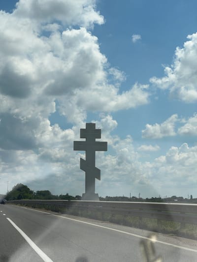 The image shows an Orthodox cross silhouetted against a cloudy sky. The cross is large and white, with a distinctly Eastern Orthodox design. The scene appears to be a roadside, with a guardrail and asphalt road visible on the right side of the frame. Trees and distant structures are visible on the horizon beyond the guardrail, suggesting a rural or semi-rural setting. The sky is filled with dramatic, white cumulus clouds against a bright blue backdrop, indicating it is daytime and likely fair weather despite the clouds. The orientation of the image is portrait, and the composition places the cross prominently in the upper portion of the frame, partially obscured by clouds. The image was captured in Yelizavetinskaya, Russia. There is no text visible in the image.