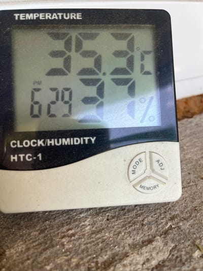 This is a close-up, eye-level shot of a digital thermometer and hygrometer resting on a rough, textured surface, likely concrete or stone. The device is black with a digital display that shows two readings. The left side of the display shows "TEMPERATURE" vertically oriented, with the current temperature reading of "74.7" degrees Fahrenheit. To the right of the temperature reading, there is a symbol for percent, indicating humidity, which is currently reading "62.9" percent.

The right side of the device is white and features three circular buttons arranged in a triangle: "ADJ," "MODE," and "MEMORY." The device is labeled "CLOCK/HUMIDITY HTC-1" vertically along the right edge.

The background is out of focus, showing the textured surface on which the device sits, with hints of organic material, possibly soil or leaf litter, suggesting an outdoor setting. The lighting suggests it is daytime, but the specific time is not discernible. There are no people or animals visible in the image. The device appears slightly dusty, indicating it is in use in an environment where such particles are present. The overall scene is simple, focusing on the functionality of the weather monitoring device.