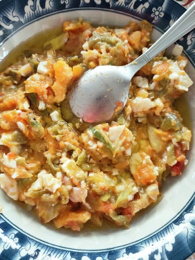 A close-up, overhead shot shows a partially eaten dish of scrambled eggs with vegetables in a blue and white patterned ceramic bowl. A silver spoon is placed in the dish. The dish appears to be a savory meal, with chopped red peppers, green peppers, and pieces of scrambled egg mixed together in a light oil. The bowl is white with a decorative blue pattern along the rim. The image captures a simple meal, possibly prepared at home. There is no specific indication of the city of Ariana, Tunisia in the image itself.