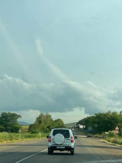 The image shows a slightly overcast day on a rural road in Tatarka, Russia. The perspective is from inside a vehicle, looking forward and slightly to the right. The primary subject in the foreground is the rear of a white SUV with a spare tire mounted on the tailgate. The license plate is visible and reads "K428KB 125". The road is asphalt and has a white line marking the lane.
To the right of the road, there are lush green trees and vegetation, with a few wildflowers visible. In the distance, more trees and a hillside can be seen. There are several traffic signs visible along the road. One is a triangular sign with two red arrows indicating a fork in the road. Another sign further down the road is blue and rectangular with text that appears to be in Cyrillic. There are other vehicles on the road ahead, including a dark-colored car and what appears to be a small bus or van. Billboards are also present along the side of the road, one of which has an image of a person.
The sky is filled with large, somewhat dramatic clouds, suggesting changeable weather. The overall impression is one of travel through a scenic, rural landscape. The time of day appears to be daytime, likely late morning or afternoon, given the light conditions. There are no people directly visible in the frame.