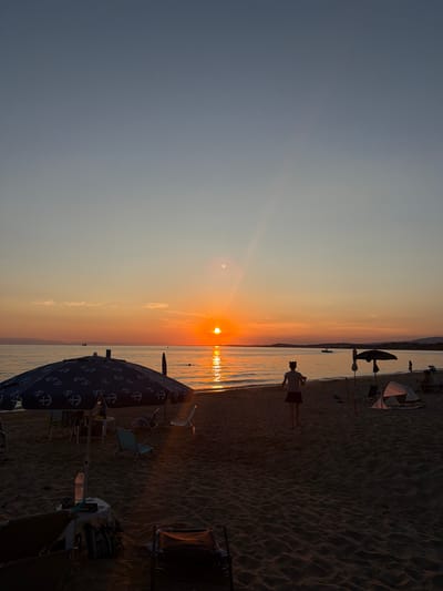 The image depicts a tranquil sunset scene on a sandy beach in Πόρτες, Greece. The sun, a vibrant orange orb, is setting over the calm, reflective water of the sea, casting a warm glow across the sky and water. The sky transitions from a pale blue at the top to a warm gradient of yellow, orange, and pink near the horizon. Several umbrellas are set up on the sand, providing shade for beachgoers. One large umbrella in the foreground features a pattern of white anchors on a dark blue background. Beach chairs and a small table are visible beneath the umbrella, suggesting a relaxed atmosphere. In the middle ground, a person is walking away from the camera towards the water, their silhouette outlined against the sunset. Further down the beach, other individuals can be seen, some sitting under umbrellas or near small tents. Boats are scattered on the water, adding to the sense of a peaceful evening by the sea. The sand is textured with ripples from the wind, and shadows are cast by the setting sun.