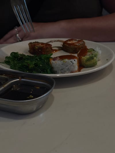 A close-up view of a dining table in Boca Raton, United States, features a white, circular plate holding multiple food items. On the plate, three pieces of golden-brown, possibly fried, cylindrical items are topped with a reddish-orange sauce. To their left, a mound of dark green seaweed salad is present. In the lower part of the plate, two sushi rolls are visible: one composed primarily of white rice with a reddish top and a light orange sauce drizzle, and another with an exterior of sliced green avocado, also topped with reddish sauce. In the immediate foreground on the left, a multi-compartment metallic dish contains a dark, reflective liquid, likely a dipping sauce, with small, light-colored particles floating within. Above the plate, a human hand, specifically a left hand adorned with a silver ring on the ring finger and another on the middle finger, holds a four-tined silver fork, positioned as if to select food. The person's forearms and a dark-colored garment are visible in the blurred background. The setting appears to be an indoor dining establishment.