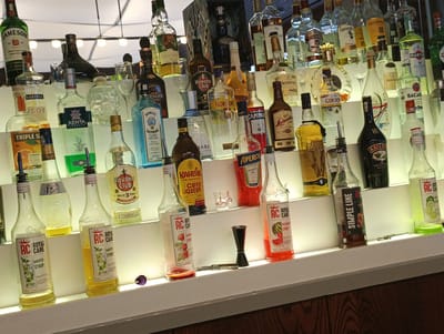 The image depicts a well-stocked bar shelf with numerous bottles of various alcoholic beverages and syrups. The bottles are arranged on multiple tiers, illuminated from below, creating a visually appealing display. Prominent brands visible include Jameson, Havana Club, Gordon's, Aperol, Martini, Baileys, and several bottles of "Royal Cane" syrup in flavors like Mojito, Strawberry, and Watermelon. There are also some unidentifiable bottles and bar tools like a jigger. The background suggests an indoor setting, possibly a bar or restaurant, with decorative lighting fixtures. The overall scene is clean and orderly, showcasing a selection of popular liquors and mixers. The location is stated to be Buzovna, Azerbaijan.