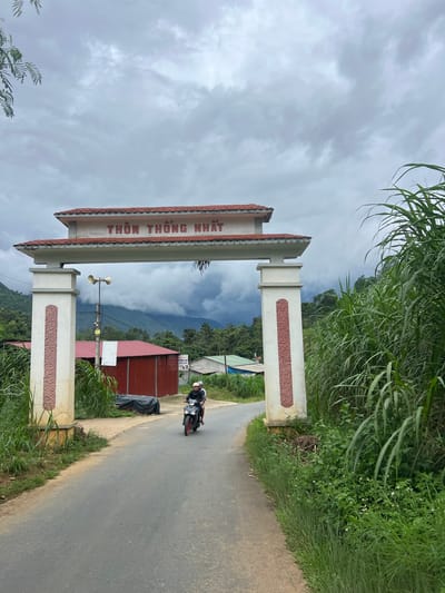 The image shows an archway with Vietnamese writing that reads "THÔM THỐNG NHẤT" over a paved road leading through it. The archway is white with red tile roofing and decorative red panels on its support pillars. A person is riding a scooter away from the camera through the archway. The road is flanked by tall green grass and foliage. In the background, there are some red-roofed buildings, hills covered in trees, and a cloudy sky. The scene appears to be an entrance to a village or community.