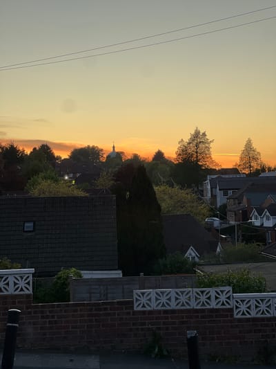 This media file captures a serene sunset or sunrise over a residential area in High Wycombe, United Kingdom. The image is dominated by the sky, which transitions from a brilliant orange and yellow along the horizon on the left to a cooler, hazy blue-grey in the upper right. Two dark, thin power lines traverse the upper left portion of the sky.
A dense band of trees and foliage forms a prominent silhouette across the middle ground, obscuring parts of the landscape below. Interspersed among the trees and visible above the horizon are several residential houses, their pitched roofs and upper stories dimly lit or silhouetted against the sky. A distinctive architectural detail is a distant domed building, topped with a spire, which stands out against the vibrant sky, possibly hinting at a local landmark such as the Guildhall cupola.
In the foreground, on the right side of the image, a decorative white concrete block fence with an open, repeating circular and star-like pattern is visible. Below this, a section of a red brick wall continues, and to its immediate right, a dark vertical pole, likely a streetlamp or utility pole, stands beside what appears to be a road or pavement.
The time of day is clearly either sunset or sunrise, creating a dramatic and peaceful atmosphere with strong contrasts between the bright sky and the darkened landscape elements. The weather appears clear and calm. There are no people or discernible activities taking place, and no visible text in the image.