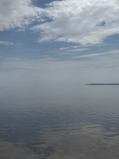 The image depicts a body of water, possibly a lake or bay, with a narrow strip of land visible in the distance. The sky is filled with clouds, and their reflection can be seen on the water's surface. The water is rippled, creating a textured pattern. The image is taken from a slightly elevated position, looking out over the water. The location could be near Şereflikoçhisar, Turkey, as it is near Lake Tuz, but the exact location cannot be determined from this image.